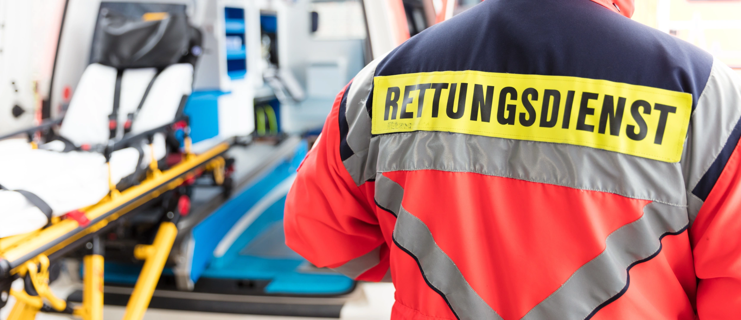 Rettungssanitäter steht mit Rettungsdienst beschrifteter Jacke auf dem Rücken neben einem Krankenwagen. Eine Krankentrage ist am geöffneten Krankenwagen ersichtlich.